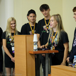 Grupa młodzieży przemawia zza mównicy. Wszyscy ubrani są w czarne koszulki z pikselowym logo województwa