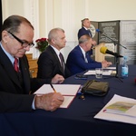 Czterech mężczyzn, trzech siedzi za stołem, jeden przemawia zza mównicy