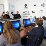 Kilka osób przed komputerami obserwuje trzyosobową grupę mężczyzn, jeden z nich prezentuje sprzęt elektroniczny
