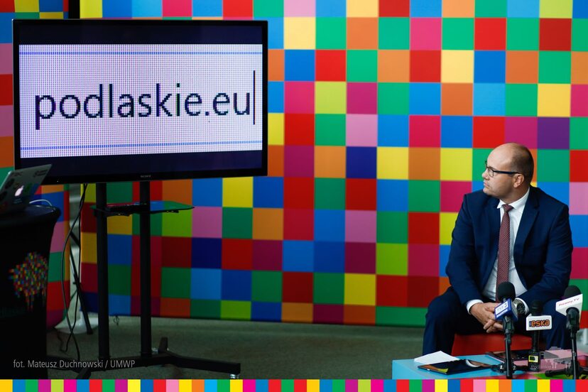 Marszałek Artur Kosicki siedzący po prawej i wpatrujący się w lewą stronę, gdzie na stojaku jest monitor, na którym wyświetlany jest napis podlaskie.eu