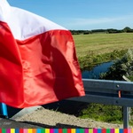 Na tle pola i rzeki powiewająca flaga biało-czerwona.