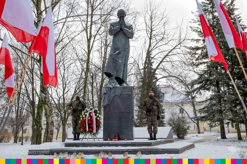 Na zdjęciu widoczny pomnik bł. ks. J. Popiełuszki i stojący żołnierze. Pod monumentem widoczne znicze oraz wieniec. Obok widoczne biało-czerwone flagi.