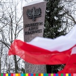 Biało-czerwona flaga na tle pomniku z metalowym orłem i napisem Żołnierzom Armii Krajowej.