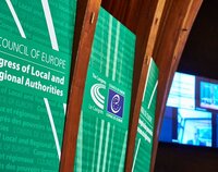 Po lewej zielone plansze z napisem w języku angielskim: Kongres Władz Lokalnych i Regionalnych Europy. Po prawej ekran monitora z uczestnikami.