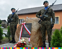 Dwóch żołnierzy stojących przy głazie z tablicą pamiątkową. Tablica przewiązana biało-czerwoną wstęgą