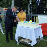 Mężczyzna kroi tort w obecności dwóch kobiet. W tle widoczna scena