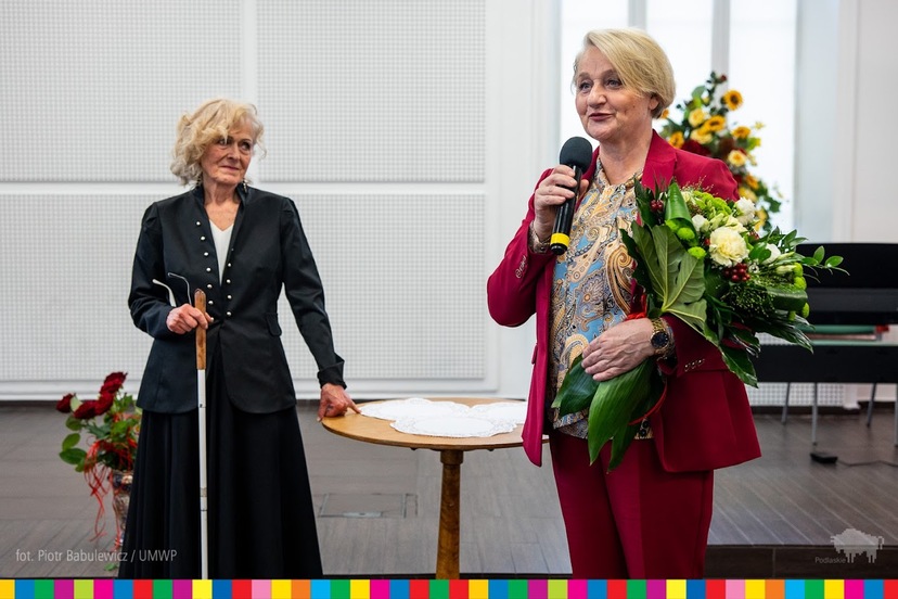 Wiesława Burnos trzyma w ręku kwiaty i przemawia przez mikrofon. Obok niej stoi kobieta