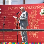 Mężczyzna przemawia na tle czerwonej ścianki z napisami Powiat Łomżyński