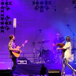 Kobieta i mężczyzna z gitarą na scenie, w tle widać innego mężczyznę grającego na perkusji 