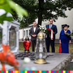 Przed pomnikiem stoi trzech mężczyzn i kobieta w kapeluszu. Dwóch mężczyzn trzyma wiązanki kwiatów w kolorach biało-czerwonych.