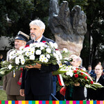 Mężczyzna w garniturze trzyma kwiaty wkolorach biało-czerwonych, po lewej stoi mężczyzna w mundurze, za nimi uczestnicy wydarzenia. 