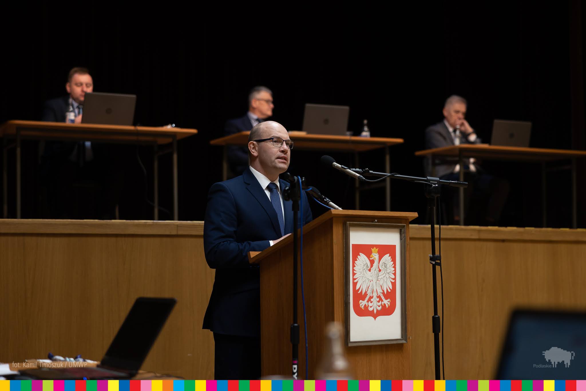 Marszałek Artur Kosicki zabiera głos na mównicy z godłem Polski. W tle za nim widać trzech siedzących meżczyzn