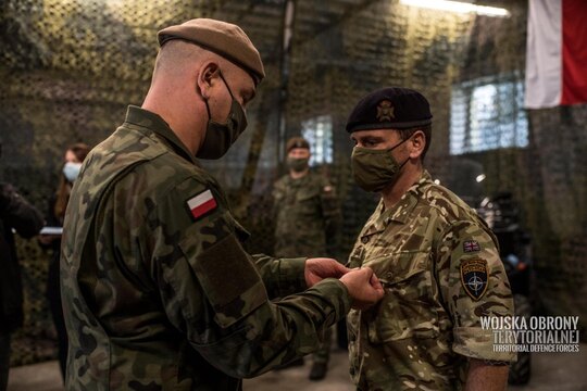 Żołnierz w polskim mundurze przypina order stojącemu obok żołnierzowi. W tle inne osoby w mundurach.