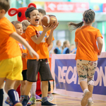 Zadowolony chłopiec w pomarańczowej koszulce chwyta piłkę futbolową. Obok niego biegające dzieci.