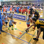 Na pierwszym planie zawodnicy Lowlanders Białystok ćwiczą z grupą dzieci rzuty piłką futbolową. Na drugim planie dzieci w pomarańczowych koszulkach. W tle widownia.