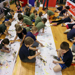Grupa kilkunastu przedszkolaków rysuje na kartkach rozłożonych na parkiecie. Widok z góry.