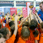 Dzieci w pomarańczowych koszulkach wyciągają ręce w górę, aby dotknąć pucharu. Po prawej instruktor Przedszkoliady w niebieskim stroju. 