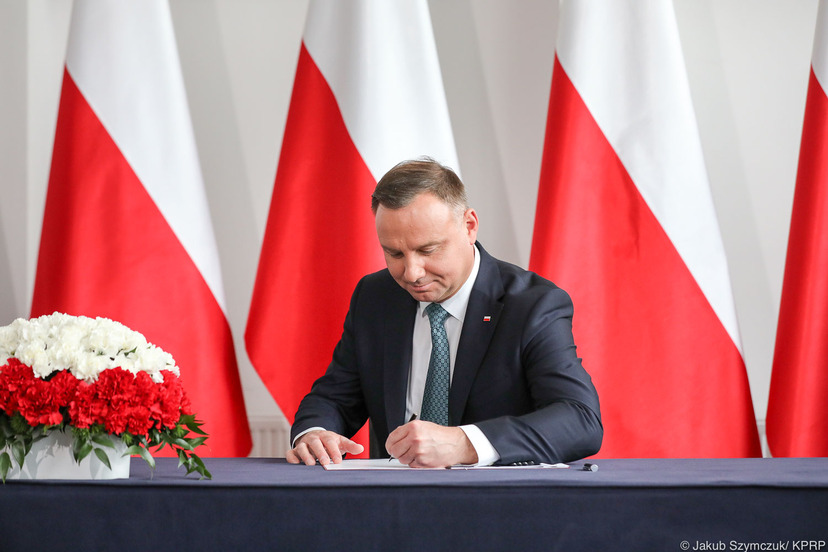 Prezydent RP podpisuje ustawę przy stole na tle polskich flag