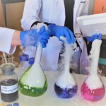 Trzy laboratoryjne szklane pojemniki z kolorową dymiąca cieczą, które trzymają osoby w rękawiczkach i białych fartuchach