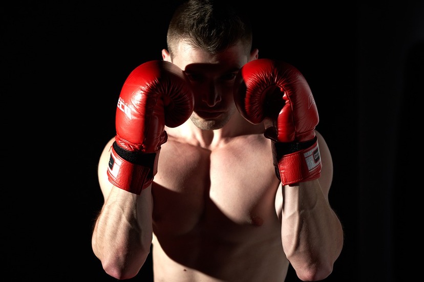 Bokser stojący na czarnym tle, jego twarz spowita w cieniu rzucanym przez czerwone rękawice bokserskie