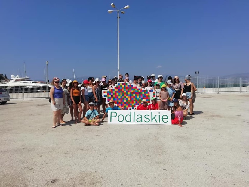 Grupa ludzi na plaży, środkowe osoby przytrzymują wielkiego żubra - logo Województwa Podlaskiego z napisem: Podlaskie.