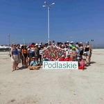 Grupa ludzi na plaży, środkowe osoby przytrzymują wielkiego żubra - logo Województwa Podlaskiego z napisem: Podlaskie.