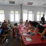 Dzieci siedzące przy stołach zasłanych foliowymi obrusami, pod opieką kilku dorosłych uczą się tkania. 