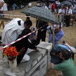 Ksiądz oraz mężczyzna wkładają kamień węgielny w fasadę budynku. Dookoła znajduje się dużo osób uczestniczących w wydarzeniu.