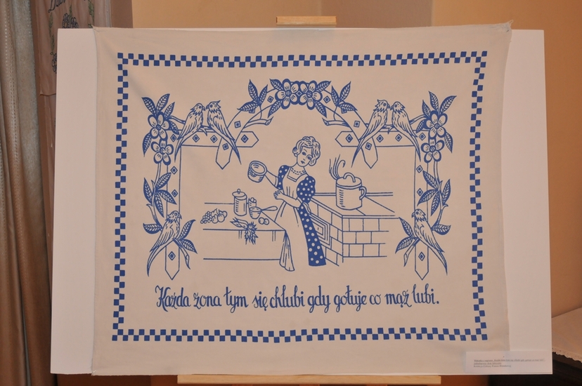 Biała makatka z niebieskim wizerunkiem kobiety w kuchni i napisem Każda żona tym się chlubi gdy gotuje co mąż lubi.