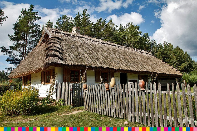 drewniany dom kryty słomą obok którego jest drewniany płot z glinianymi garnkami na sztachetach.