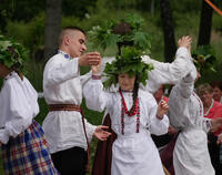 Tańczy mężczyzna i cztery kobiety z zielonymi wieńcami na głowach. Wszyscy  ubrani w białe stroje ludowe.