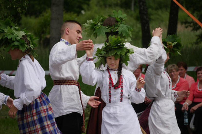 Tańczy mężczyzna i cztery kobiety z zielonymi wieńcami na głowach. Wszyscy  ubrani w białe stroje ludowe.