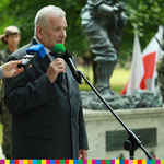 Starszy człowiek przemawiający pod pomnikiem na tle biało-czerownych flag