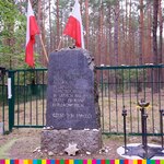 Kamienny pomnik z tablicą z flagami biało czerwonymi w tle oraz ogrodzenie cmentarza
