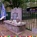 Na pierwszym planie jest widoczny pomnik z tablicą upamiętniającą Zagładę Żydów Tykocińskich z flagą w tle