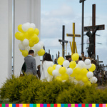 Kilkoro osób trzymających ogromne pęki białych i żółtych balonów, stojąca na tle krzyży 