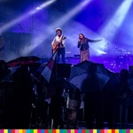Mężczyzna i kobieta śpiewający na scenie oraz publiczność stojąca pod parasolami