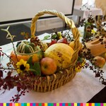 Na stole przykrytym białym obrusem stoi ozdobny kosz z warzywami Obok koszyka  leżą warzywa..