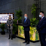 Na zdjęciu cztery osoby stoją. W tle rośliny ustawione na podestach oraz ekran z treścią