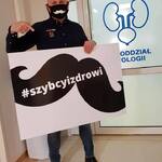 Jarosław Kazberuk trzyma tablicę z wizerunkiem czarnych wąsów i hasztagiem #szybcyizdrowi.