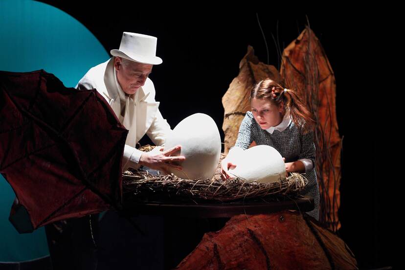 Mężczyzna w białym fraku i cylindrze trzyma w dłoniach wielkie kacze jajo, obok dziewczynka trzyma drugie