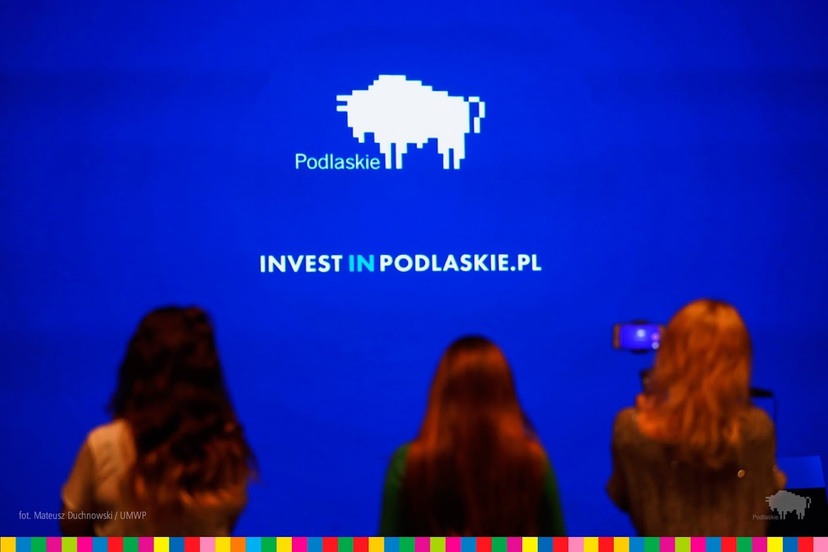 Niebieska plansza z napisem InwestinPodlaskie i logo województwa. Przed nią trzy ludzkie sylwetki