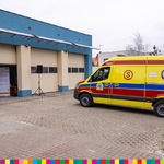 Widok ambulansu w barwie żółtej, czerwonej, niebieskiej stojącego przed garażami