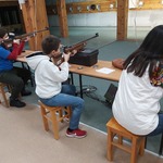 Trzy młode osoby trzymające broń strzelecką siedzą na stanowiskach strzelniczych.