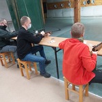 Trzech mężczyzn  trzymających broń strzelecką siedzi na stanowiskach strzelniczych.