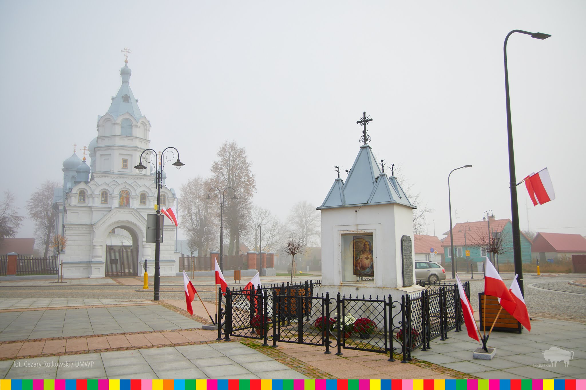 Kapliczka z obrazem Jezusa i biało - czerwonymi flagami. Na drugim planie cerkiew Świętych Apostołow Piotra i Pawła w Wasikowie.