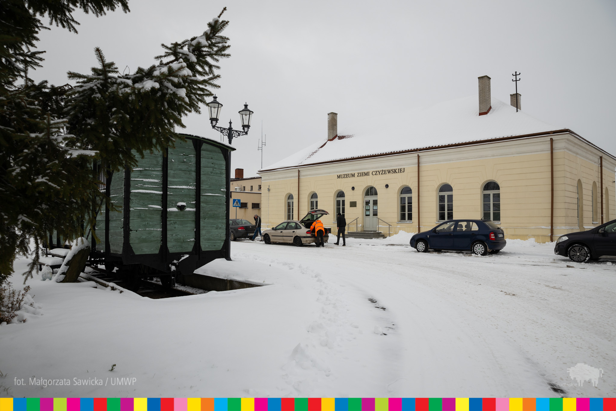 Budynek Muzeum Ziemi Czyżewskiej. Przed budynkiem stoją samochody. Obok stoi drewniany wagon