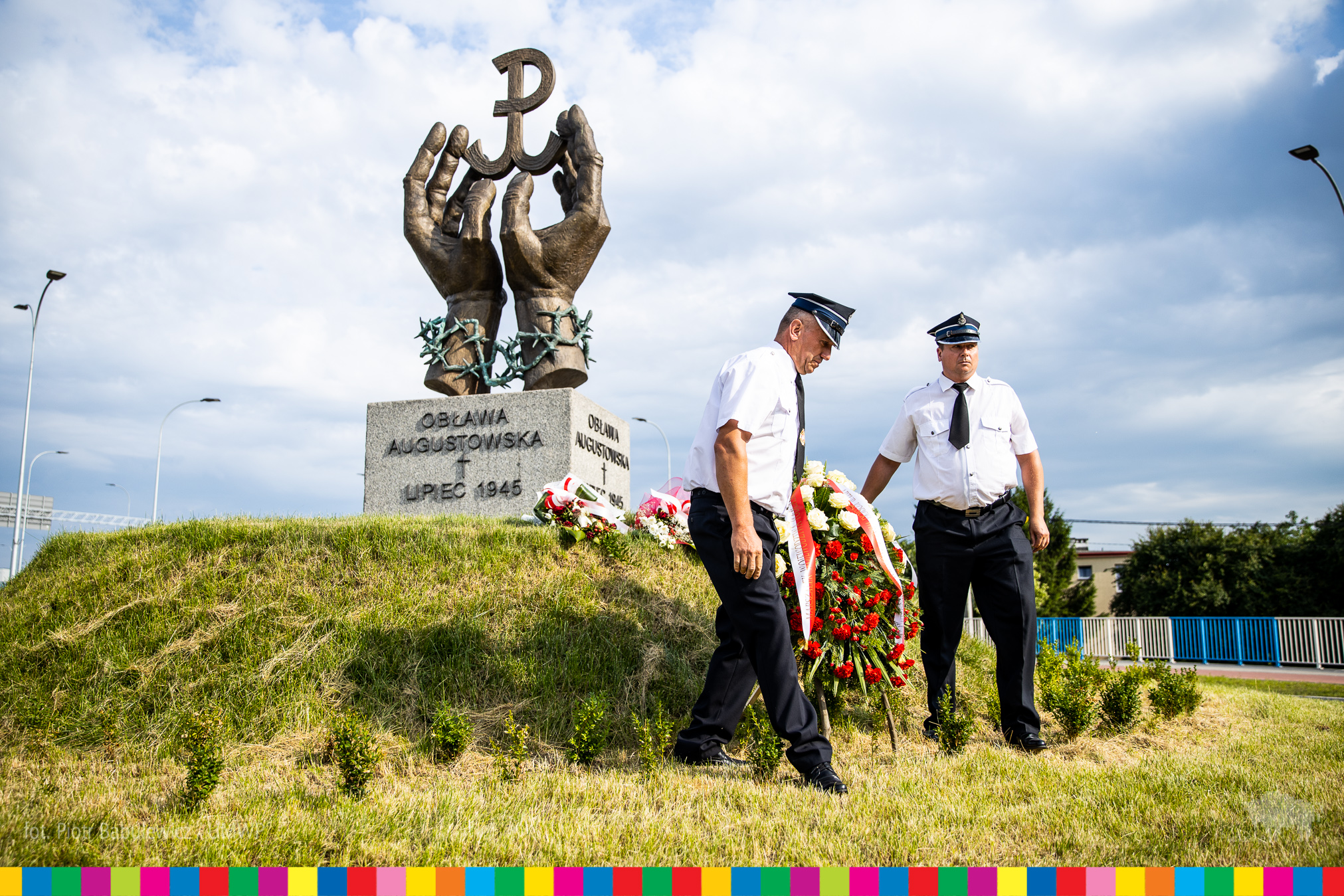 Pomnik w kształcie dwóch dłoni trzymających literę P. Dwóch strażaków wnosi pod niego wieniec. 