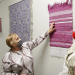 Dwie kobiety w maseczkach oglądające i dotykające wiszącą na ścianie tkaninę