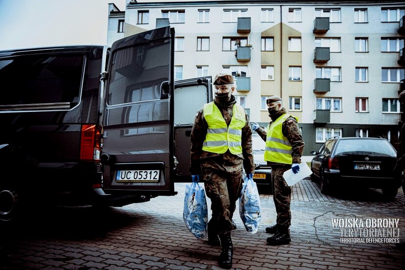 Dwóch żołnierzy wynoszą torby z pojazdu wojskowego. Na drugim planie blok mieszkalny.